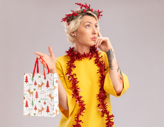 思考体贴的金发女郎头戴圣诞花环 脖子上戴着金箔花环 手里拿着圣诞礼品袋 一边看一边做着思考的动作 在白色的背景上与世隔绝包周围礼物