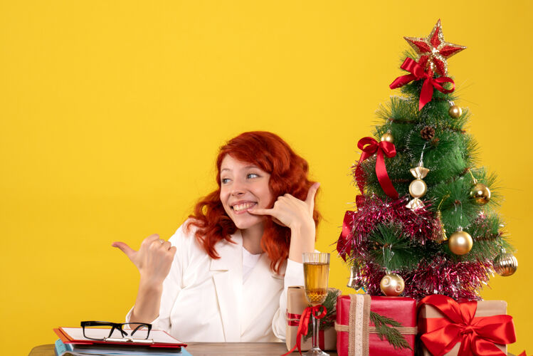 安排女工人坐在桌子后面 手里拿着圣诞礼物 树在黄色上微笑树情感工人