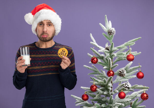 看着戴着圣诞帽的悲伤的白人年轻人站在装饰好的圣诞树旁 手里拿着一杯牛奶和饼干 看着隔离在紫色背景上的摄像机玻璃牛奶帽子