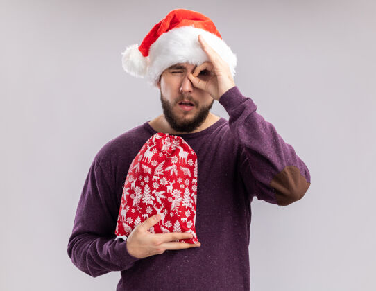 ”穿着紫色毛衣 戴着圣诞帽的年轻人手里拿着红包 手里拿着礼物 看着白色背景上的这个牌子唱着“ok