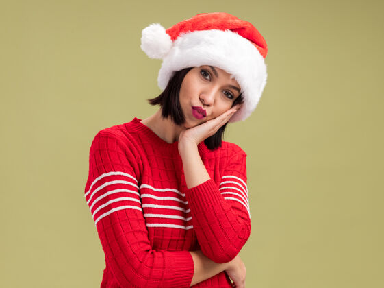 帽子困惑的年轻女孩戴着圣诞帽看着相机 手放在脸上 噘着嘴唇 在橄榄绿的背景上 有复制空间保持噘嘴脸