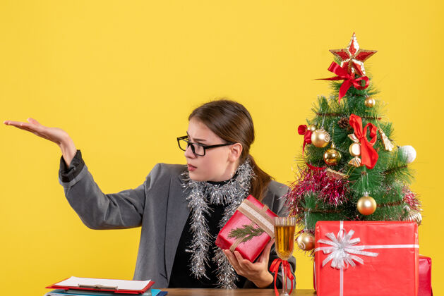 笔记本电脑前视图年轻女孩戴着眼镜坐在桌旁 展示圣诞树和鸡尾酒礼物年轻人展示树