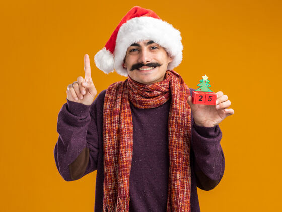 温暖快乐的小胡子男人戴着圣诞老人帽 脖子上围着暖和的围巾 手里拿着玩具方块 上面写着日期25 他笑得很开心 食指站在橙色的背景上举行高兴脖子
