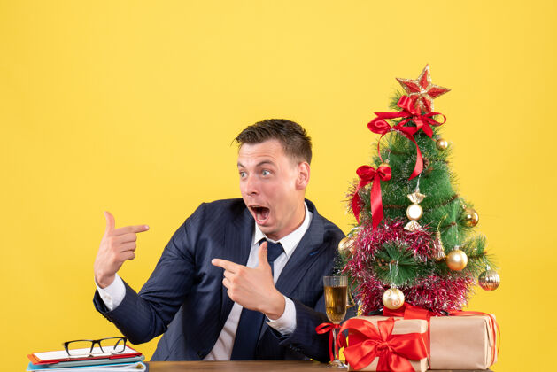 人前视图惊奇的人指着自己坐在圣诞树旁的桌子和黄色墙上的礼物他自己桌子男