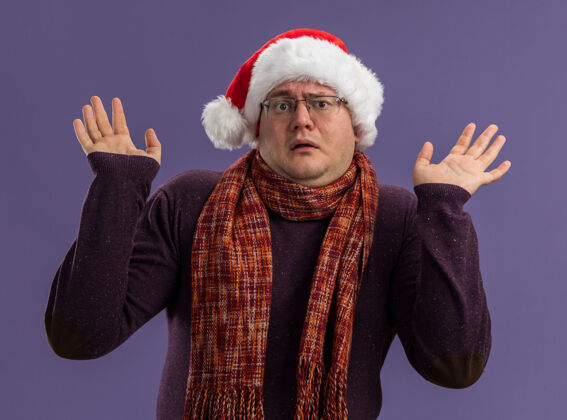 空笨手笨脚的成年男子戴着眼镜 戴着圣诞帽 脖子上围着围巾 看着镜头 紫色背景上孤立着空空的双手圣诞老人周围不知所措