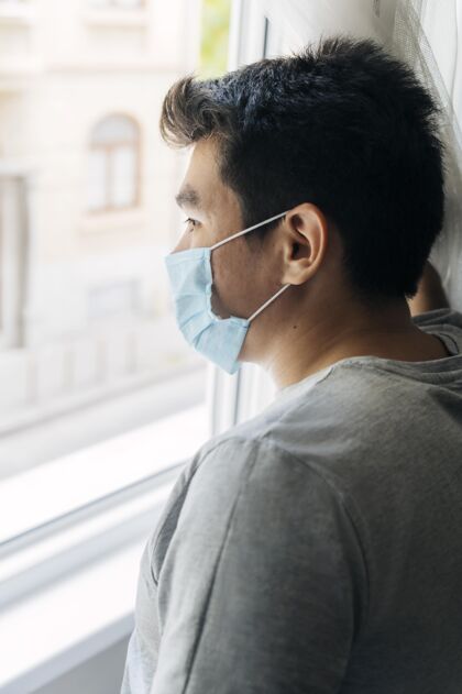 面罩大流行期间 家里戴着医用口罩的男人透过窗户看医学面罩医学男性