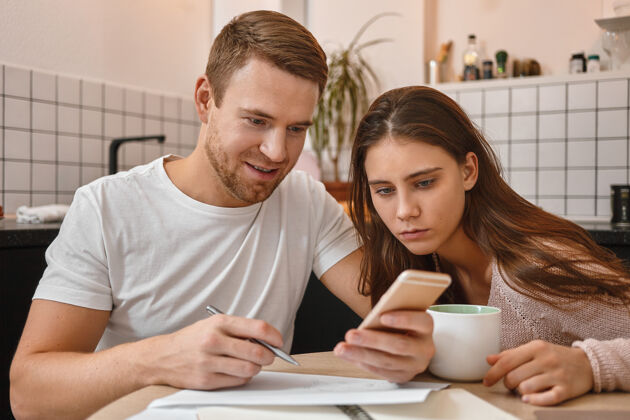 关系好奇的年轻女子坐在厨房的餐桌旁 在丈夫手中的手机上读着积极的消息迷人的男子正在填写金融文件 通过手机使用网上银行应用程序付款预算不开心