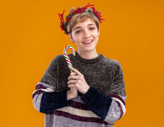头快乐的年轻漂亮女孩戴着圣诞花环 手持圣诞甜甜的手杖孤立在橙色的墙上快乐手杖漂亮