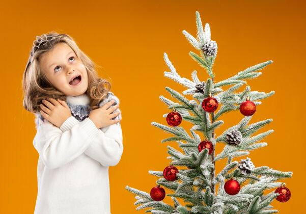 圣诞节兴奋地抬头望着站在圣诞树旁的小女孩 戴着头饰 脖子上戴着花环 手放在肩膀上 隔离在橙色的墙上小穿站着