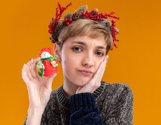 雕像困惑的年轻漂亮女孩戴着圣诞花环 手持小圣诞雪人雕像 手放在脸上 隔离在橙色的墙上圣诞节花环漂亮