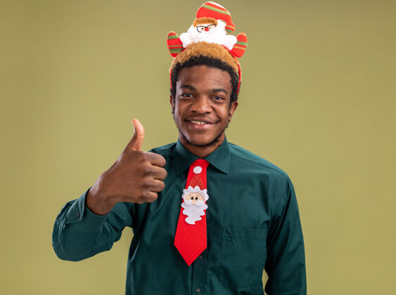 微笑快乐的非洲裔美国人 戴着有趣的圣诞老人戒指 打着红色领带 站在绿色的背景下 看着镜头 竖起大拇指微笑着非洲站着搞笑