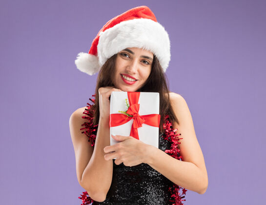 女孩微笑着的年轻漂亮女孩戴着圣诞帽 脖子上戴着花环 手里拿着紫色背景上孤立的礼盒帽子抱着盒子