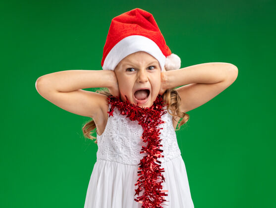 戴恼怒的小女孩戴着圣诞帽 脖子上戴着花环 耳朵被隔离在绿色的墙上帽子盖圣诞节