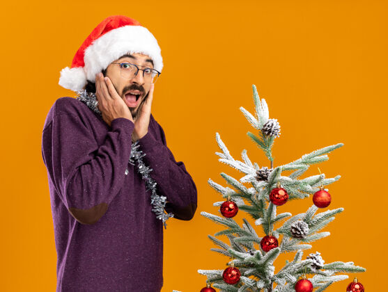 戴着兴奋的年轻帅哥站在圣诞树旁 戴着圣诞帽 脖子上戴着花环 手放在橘色的墙上孤立的脸颊上旁边脖子花环