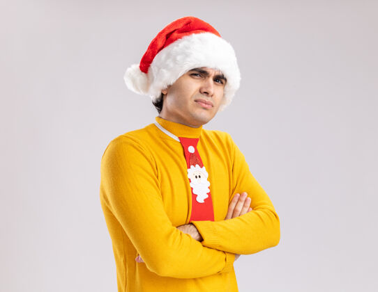 怀疑穿着黄色高领毛衣 戴着圣诞帽 打着滑稽领带的年轻人抱着怀疑的表情看着摄像机 双臂交叉站在白色背景上高领毛衣手臂帽子