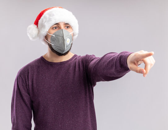保护一个穿着紫色毛衣 戴着圣诞老人帽 戴着护面面具的年轻人 一边焦急地用食指指着白色背景上的东西指数手指指着