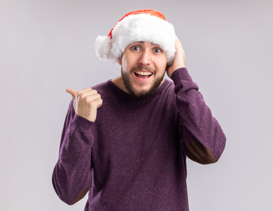 高兴穿着紫色毛衣 戴着圣诞老人帽的快乐的年轻人看着摄像机 愉快地微笑着 拇指背站在白色的背景上帽子拇指微笑