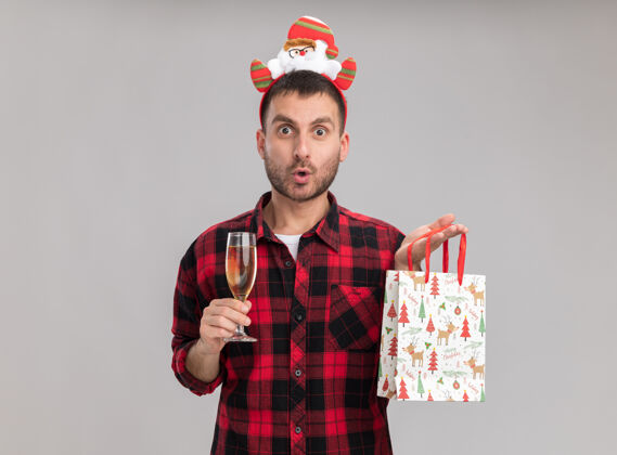 男人令人印象深刻的年轻白种人戴着圣诞头带 拿着圣诞礼品袋和一杯香槟 隔离在白色墙壁上 留有复制空间圣诞节印象深刻香槟