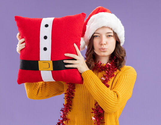 帽子自信的年轻斯拉夫女孩 戴着圣诞帽 脖子上戴着花环 抱着装饰枕头 紫色背景 有复制空间圣诞老人年轻装饰