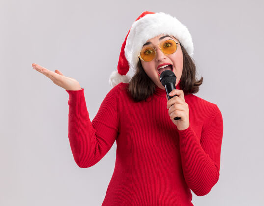 圣诞老人快乐快乐的年轻女孩 穿着红色毛衣 戴着圣诞帽 戴着眼镜 拿着麦克风 微笑着看着镜头 举起手臂站在白色的背景上微笑站着麦克风