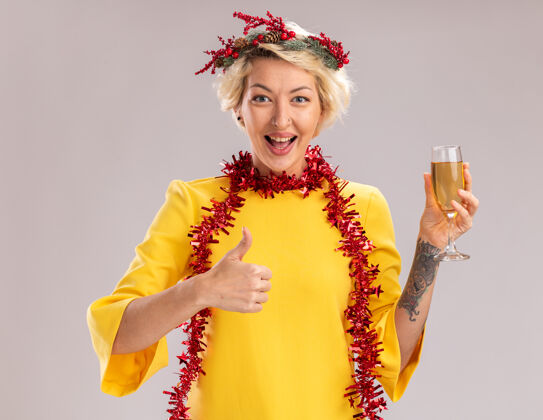 头快乐的年轻金发女人戴着圣诞花环 脖子上戴着金箔花环 手里拿着一杯香槟 在白墙上孤立地竖起大拇指周围女人拇指