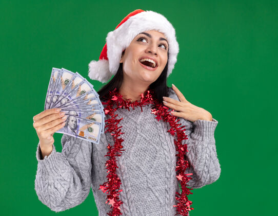帽子给人印象深刻的年轻白人女孩戴着圣诞帽 脖子上戴着金箔花环 手里拿着钱 抚摸着肩膀 孤零零地看着绿色的墙壁脖子圣诞节钱