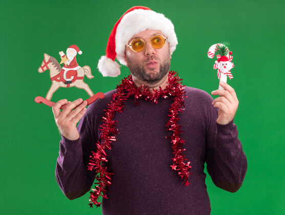 中年人给人印象深刻的中年男子戴着圣诞帽 脖子上戴着金箔花环 戴着眼镜 手里拿着糖果藤饰品和圣诞老人帽子圣诞快乐圣诞