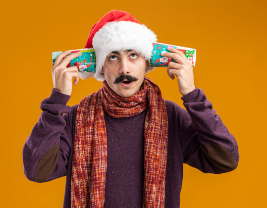 站着一个留着胡子的男人戴着圣诞老人帽 脖子上围着暖和的围巾 耳朵上戴着五颜六色的纸杯 站在橘色的背景上困惑地仰望着耳朵抱着温暖