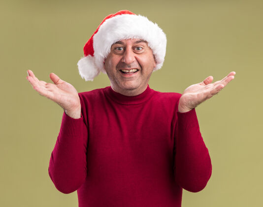 胳膊中年男子戴着圣诞老人的帽子 看着镜头 高兴而兴奋地微笑着 双臂高举 站在绿色的背景上年龄微笑圣诞节