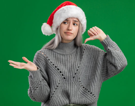 金发年轻的金发女郎穿着冬季毛衣 戴着圣诞老人帽 看起来很困惑 大拇指朝下 用手臂站在绿色背景上展示复制空间拇指站着毛衣