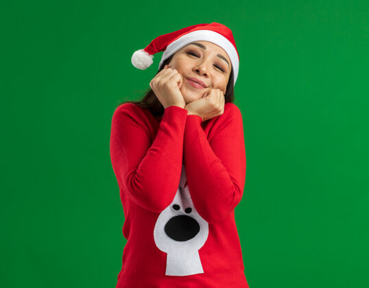 感觉快乐的年轻女子戴着圣诞老人帽 穿着红色毛衣 站在绿色的背景下 看着镜头 感受积极的情绪 微笑着圣诞节站立微笑