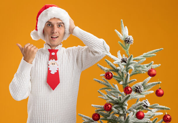 放着年轻帅哥戴着圣诞帽 打着圣诞老人的领带 站在装饰好的圣诞树旁 手放在头上 指着隔离在橙色墙上的一边年轻圣诞老人侧面