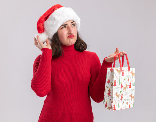 圣诞节穿着红色毛衣 戴着圣诞帽的年轻女孩 手里拿着彩色纸袋 里面装着圣诞礼物 站在白色的背景上 带着怀疑的表情抬起头来表情帽子站着