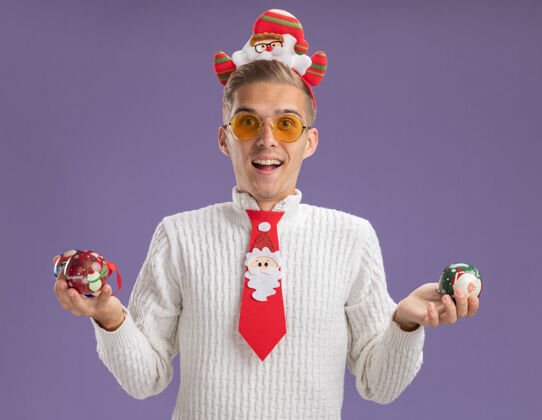 圣诞节印象深刻的年轻帅哥戴着圣诞老人的头带和领带戴着眼镜拿着圣诞球饰品孤立在紫色的墙上头带饰品圣诞老人
