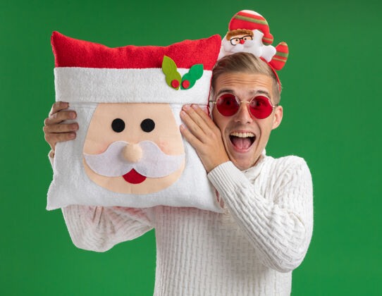 头给人印象深刻的年轻帅哥戴着圣诞老人的头带戴着眼镜抱着圣诞老人的枕头靠近头隔离在绿色的墙上枕头圣诞老人印象深刻