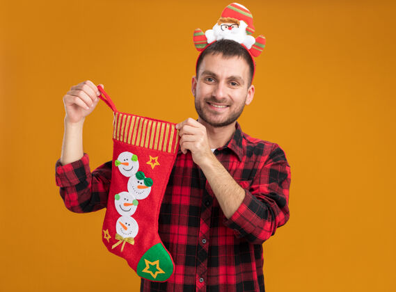 抱着微笑着的白人年轻人戴着圣诞老人的头带 手里拿着圣诞长袜 看着隔离在橙色背景上的相机 还有复制空间相机圣诞老人头带
