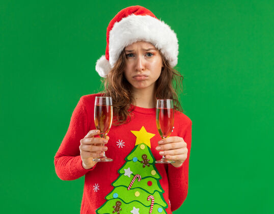 Pursing心烦意乱的年轻女子 穿着红色圣诞毛衣 戴着圣诞老人帽 手里拿着两杯香槟 神情悲伤地看着相机 噘着嘴唇站在绿色的背景上年轻毛衣嘴唇