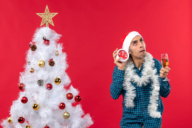 庆祝一个戴着圣诞老人帽子 拿着一杯葡萄酒 拿着一个闹钟站在圣诞树旁的困惑的年轻人圣诞老人帽子困惑