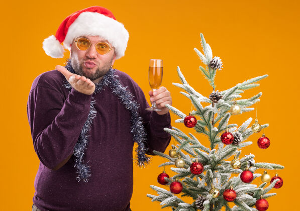 新的高兴的中年男子戴着圣诞帽 脖子上戴着金箔花环 戴着眼镜 站在装饰好的圣诞树旁 手里拿着一杯香槟举行圣诞快乐吻