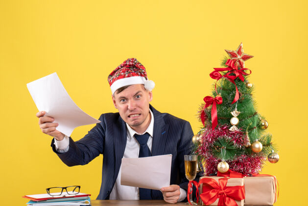 男人前视图愤怒的男子与圣诞树附近的圣诞帽坐在桌子上 并在黄色的墙上复制空间的礼物圣诞节帽子礼物