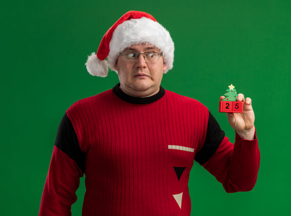 日期令人印象深刻的成年男子戴着眼镜和圣诞帽举行和看圣诞树玩具与日期隔离的绿色背景举行成人圣诞树