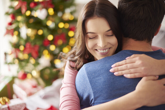 团结微笑的女人在圣诞节拥抱她的男朋友结合圣诞树拥抱