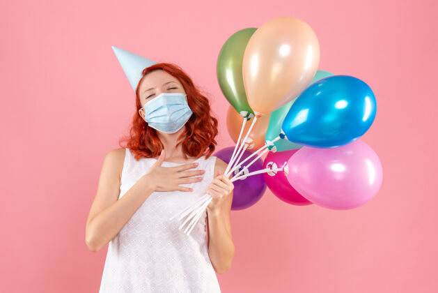 女性正面图是粉红色墙上戴着消毒面具手持彩色气球的年轻女子派对举行前面