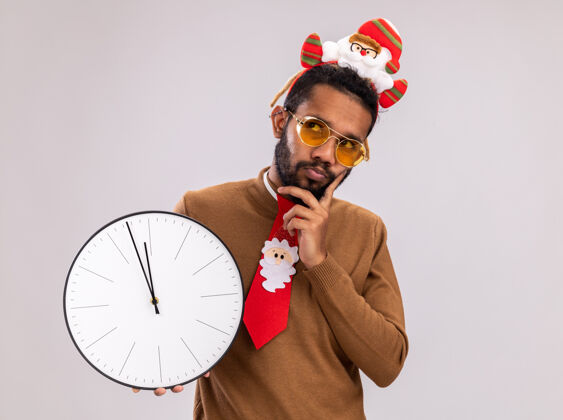 边缘一个非洲裔美国人 穿着棕色毛衣 头戴圣诞老人圈 打着有趣的红色领带 站在白色背景上 举着时钟 困惑地抬头看困惑时钟毛衣