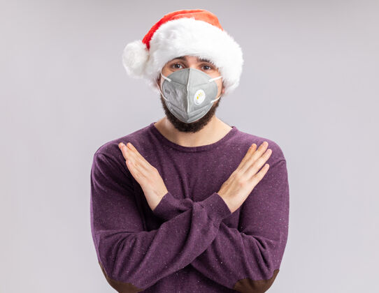 帽子一个穿着紫色毛衣 戴着圣诞帽 戴着护面面具的年轻人 严肃地看着镜头 双手交叉着脸站在白色的背景上保护圣诞老人面具