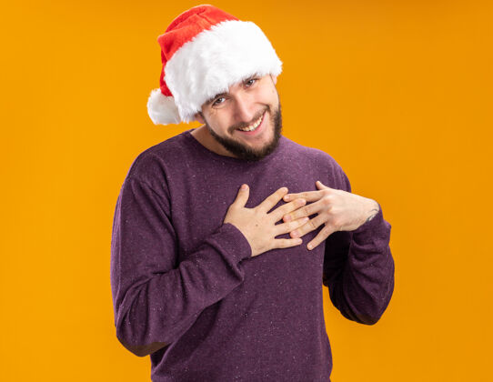 抱着穿着紫色毛衣 戴着圣诞帽的年轻人站在橙色背景下 看着相机 手放在胸前 心怀感激感觉手胸前