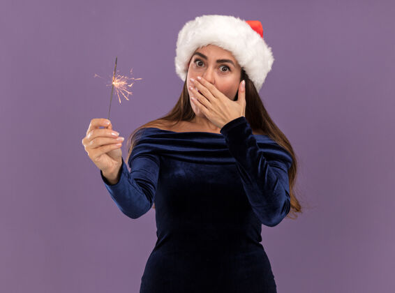 举行惊讶的年轻漂亮女孩穿着蓝色连衣裙 戴着圣诞帽 手里拿着闪闪发光的东西 嘴巴上用手隔离着紫色的背景圣诞节年轻帽子