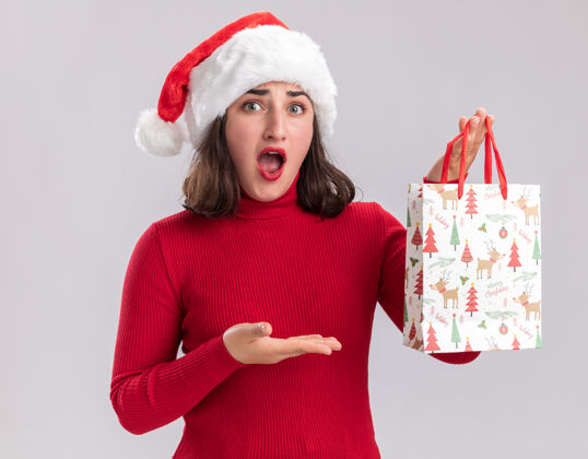 圣诞老人穿着红色毛衣 戴着圣诞帽的年轻女孩手拿着彩色纸袋 里面装着圣诞礼物 看起来很惊讶 她的手臂站在白色的背景上女孩手臂惊讶