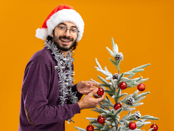脖子微笑的年轻帅哥站在圣诞树旁 戴着圣诞帽 脖子上戴着花环 橙色的背景孤立着圣诞站着微笑