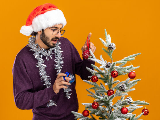 帽子想着站在圣诞树旁的年轻帅哥 戴着圣诞帽 脖子上戴着花环 手里拿着橙色背景上孤立的圣诞球小伙子花环思想